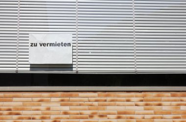 An einem Schaufenster mit heruntergelassenen, weißen Jalousien klebt ein Hinweisschild mit dem Aufdruck "zu vermieten". 