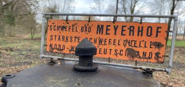 Ein alter Brunnendeckel eines Schwefelbrunnens mit einem gelben Schild auf dem der Name der Schwefelquelle steht.