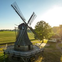 Eine Drohnenaufnahme aus der Luft, die im Vordergrund eine historische Windmühle zeigt und Felder im Hintergrund. 