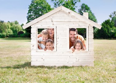 Ein kleines Gartenhaus aus Holz, das auf einem sommerlichen Rasen steht.. Aus den Fenstern schaut eine lachende Familie. 