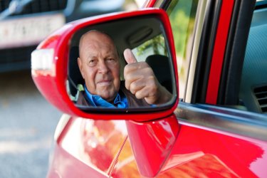 Ein älterer Mann sitzt in einem roten Auto hinter dem Lenkrad. Man sieht ihn durch den Rückspiegel in die Kamera schauen.