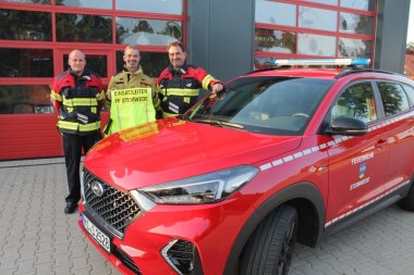 Drei Feuerwehrmänner in Montur stehen vor einem roten Garagentor an einem roten Feuerwehr-SUV.