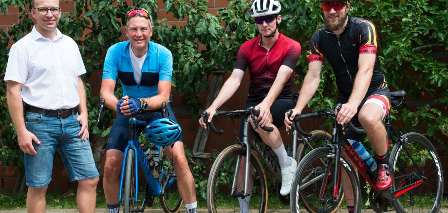 Ein Gruppenfoto mit insgesamt vier Männern. Einer davon steht links im Bild. Drei weitere Männer stehen rechts von ihm in Rennradfahrermontur an ihren Rennrädern.