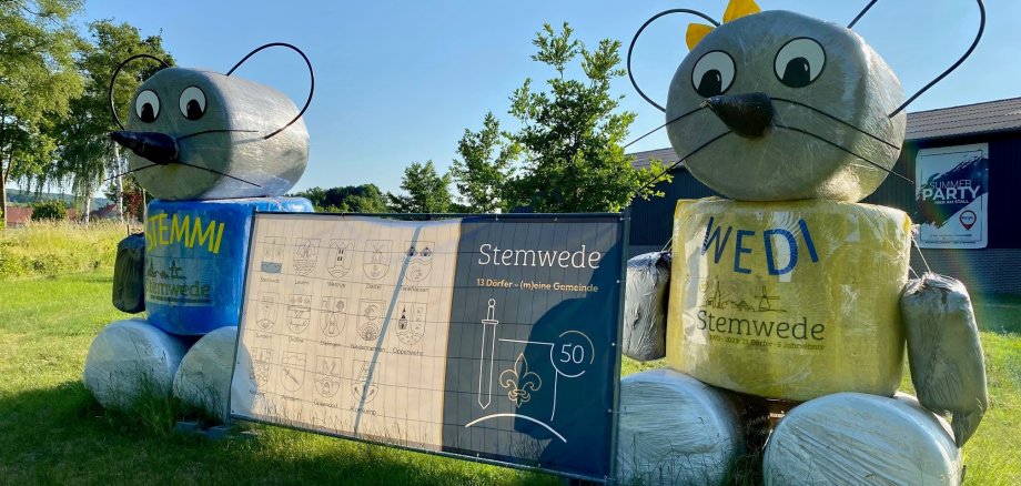 Auf einer Wiese stehen zwei Strohpuppen, die wie die beiden Maskottchenmäuse der Gemeinde Stemwede aussehen. In der Mitte der beiden Mäuse hängt ein blau-weißes Plakat, das auf das 50. Gemeindejubiläum der Gemeinde Stemwede hinweist. 