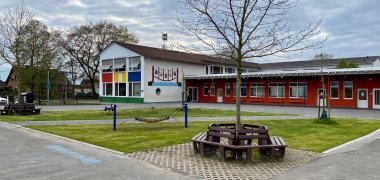 Eine kleine Rasenfläche mit Spielmodulen auf einem Schulhof. Im Hintergrund sind ein weißes und ein rotes Schulgebäude zu erkennen. 