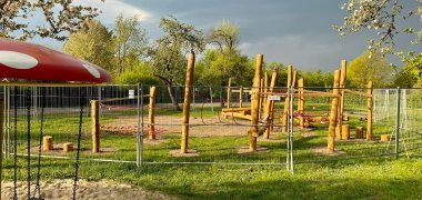 Ein eingezäuntes Spielmodul aus Holz auf einem Spielplatz. Im linken Teil ist ein Teil eines kleinen Spielplatzkarussells in Form eines Fliegenpilzes zu erkennen.