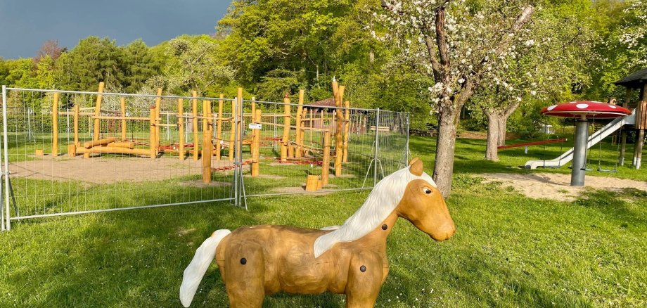 Ein Spielplatz auf dem im Vordergrund ein hölzernes Pferd zu erkennen ist und im Hintergrund ein eingezäuntes Spielmodul aus Holz. 