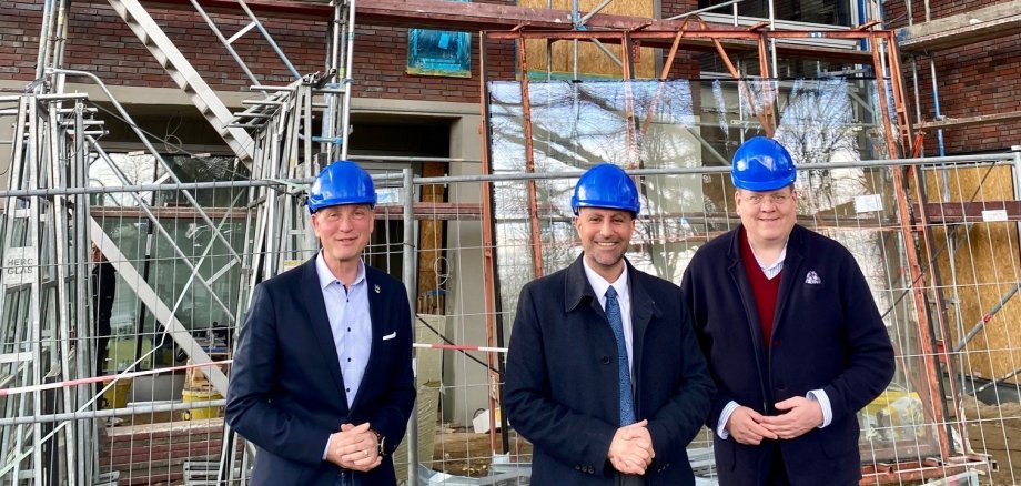 Drei Männer mit blauen Bauarbeiterhelmen stehen vor einem großen Neubau und lachen in die Kamera.