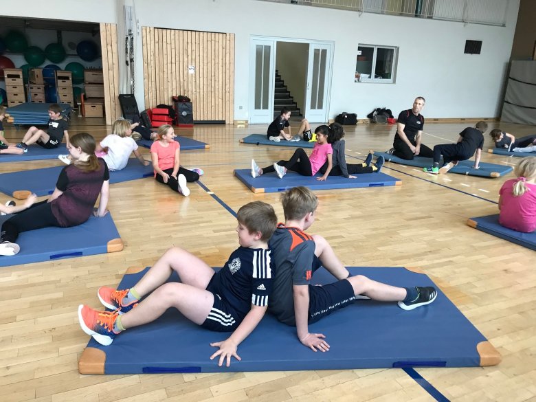 In einer Turnhalle sitzen, immer zu zweit und Rücken an Rücken, viele Kinder verteilt in einer Turnhalle. 