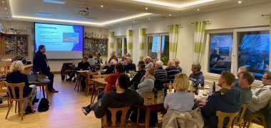 Ein Versammlungsraum, in dem etwa 30 Personen auf eine Leinwand schauen, auf der eine Präsentation gezeigt wird. 