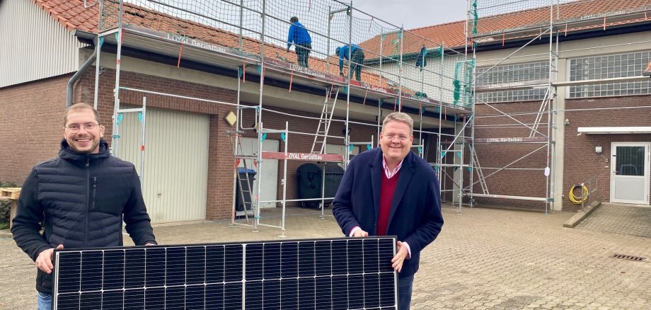 Zwei Männer, die ein Solarmodul festhalten. Im Hintergrund arbeiten Männer auf einem Gerüst, das an einem roten Gebäude steht. 