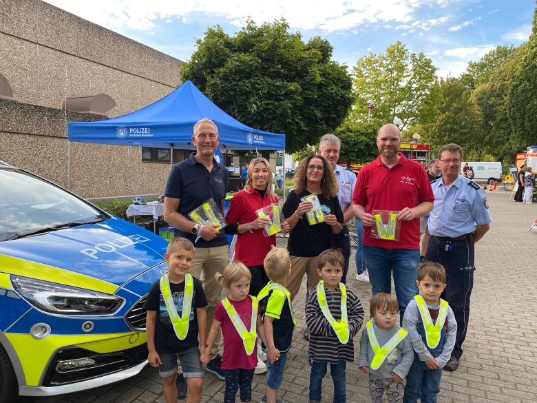 Gruppenfoto mit Kindern und Erwachsenen vor einem Polizeiauto. Die Kinder tragen reflektierende, gelbe Umhängekragen, damit sie auf dem morgendlichen Schulweg besser zu erkennen sind. 