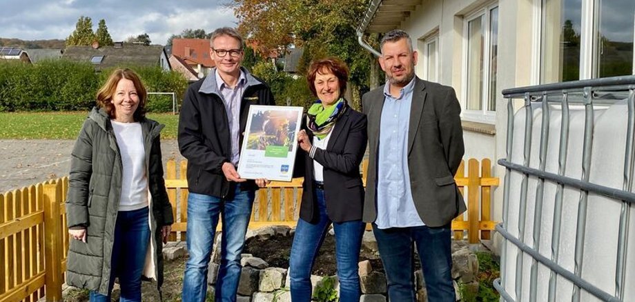 Ein Mann übergibt einer Frau eine Urkunde, während sie in einem Gemüsegarten stehen.