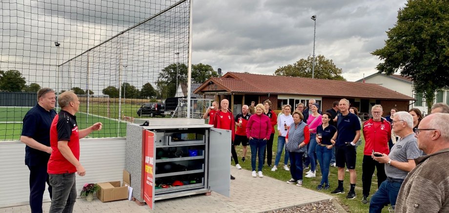 Eine Gruppe von Sportlerinnen und Sportlern, die auf einem Sportgelände vor einem geöffneten Geräteschrank steht.