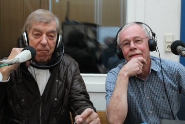 Zwei Männer sitzen in einem kleinen Tonstudio, tragen Kopfhörer und schauen in die Kamera.