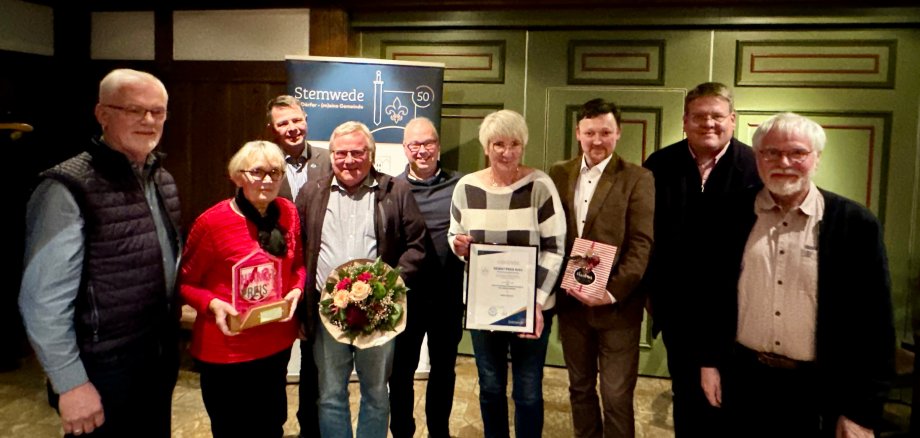 Acht Männer und zwei Frauen stehen in einem rustikalen Saal, vor einer grünen Holztür. Eine Dame hält eine Urkunde in die Kamera, die zweite Dame eine gläserne Trophäe.