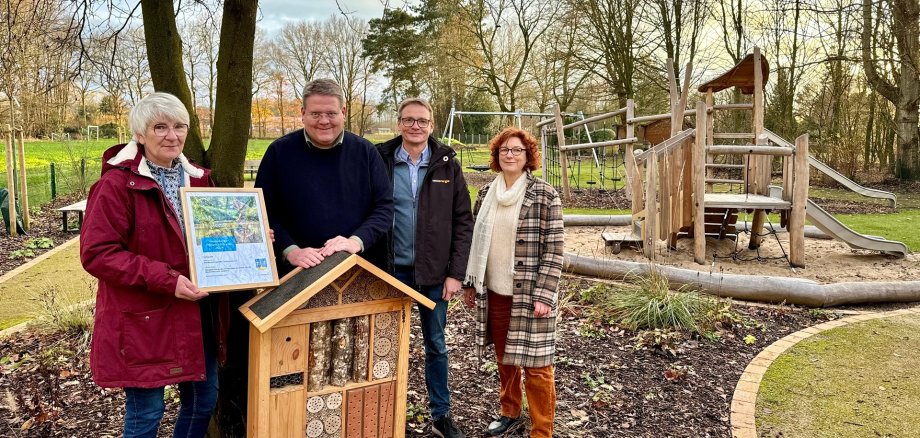 Zwei Männer und zwei Frauen stehen in einer herbstlichen Parkanlage. Ein Mann hält ein Insektenhotel, die Frau links im Bild eine eingerahmte Urkunde. Im Hintergrund ist ein Spielgerüst aus Holz zu erkennen.