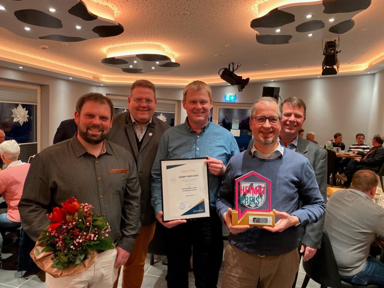 Vier Männer stehen in einem Veranstaltungsraum und lächeln in die Kamera. Einer der Männer hält einen Blumenstrauß, ein anderer eine Trophäe in der Hand.