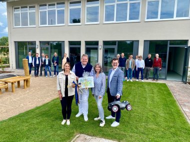 Zwei Frauen und zwei Männer stehen auf dem frisch gemähten Rasen eines Kindergarten-Außenbereichs. Sie halten ein Bobbycar sowie einen überdimensionalen Schlüssel und lachen in die Kamera. Im Hintergrund stehen eine Reihe weiterer Personen vor der Fensterfront eines Gebäudes.