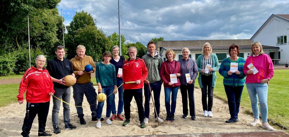 Insgesamt 12 Personen, 8 Frauen und 4 Männer, stehen auf einem Sportplatz im Sand einer Sprungkuhle und halten verschiedene Sportgeräte, die für das Sportabzeichen benötigt werden, in den Händen. Zum Beispiel Schlagbälle, Metermaß oder auch eine Harke.