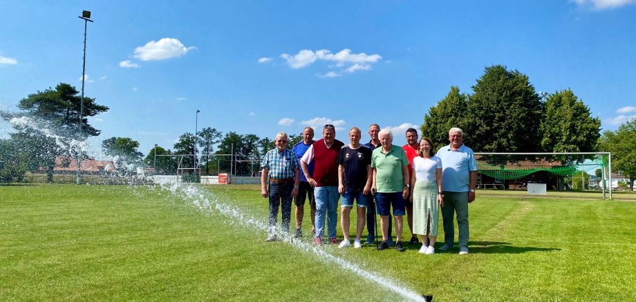 Eine Gruppe von Menschen steht an einem sonnigen Tag auf einem Sportplatz. Vor ihnen schaut eine Rasensprengerdüse aus dem Boden, die in Betrieb ist und Wasser ausstößt.