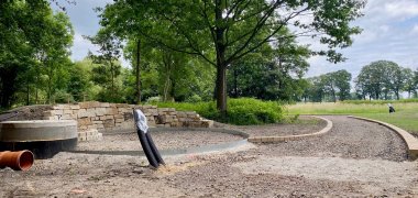 Eine Baustelle in einer parkähnlichen Landschaft. Es ist eine frisch entstandene Steinmauer zu erkennen, ein Brunnenschacht und mehrere Rohre, die aus der Erde schauen.