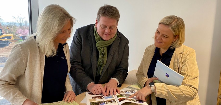 Zwei Frauen und ein Mann stehen an einem Empfangstresen und blättern gemeinsam durch eine Broschüre.