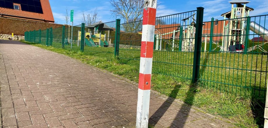 Ein rot-weiß gestreifter Absperrpoller auf einem Geh- und Radweg. Im Hntergrund ist ein Spielplatzgelände zu erkennen.