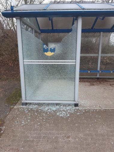 Ein gläsernes Buswartehäuschen, bei dem ein Glaselement zerstört wurde. Die Glasscheibe, auf dem zum Teil noch das Wappen der Gemeinde Stemwede zu sehen ist, ist komplett geborsten und zum Teil zersplittert.