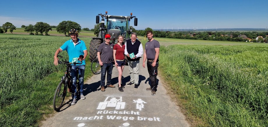 Mehrere Personen stehen bei bestem Wetter auf einem Feldweg zwischen grünen Feldern. Im Hintergrund steht ein grüner Trecker, auf der linken Seite ein Fahrradfahrer und in der MItte der Straße stehen drei Männer und eine junge Frau. Auf die Straße ist mit weißer Farbe der Schriftzug "Rücksicht macht die Wege breit" zu lesen. 