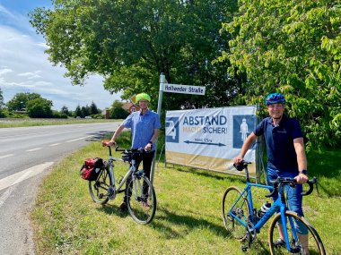 Zwei Radfahrer stehen mit Ihren Rädern vor einem großen Banner, das auf den Mindestabstand beim Überholen von Radfahrern hinweist.