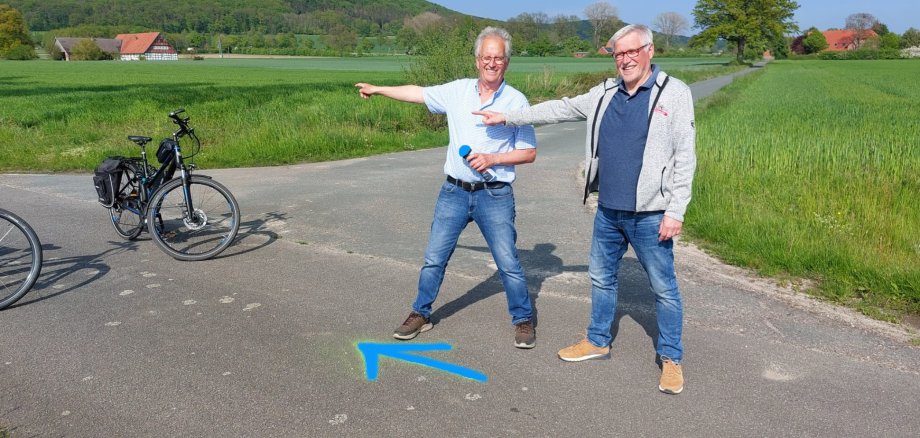 Zwei Männer stehen auf einer Straße, auf die sie gerade einen blauen Pfeil gesprüht haben. Im Bild sind auch zwei Fahrräder zu sehen.