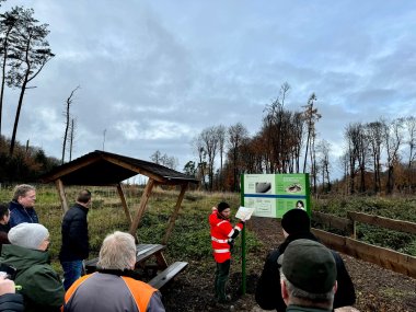 Mehrere Personen stehen an einem Hinweisschild in einem Wald. Ein Mann in einer roten Jacke scheint Erläuterungen zu den Informationen zu machen, die auf dem Schild zu lesen sind.