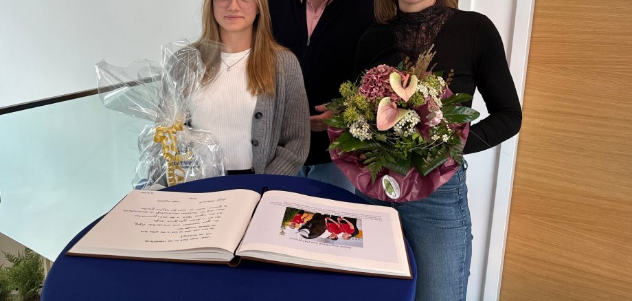 Zwei junge Frauen stehen an einem Stehtisch vor einem aufgeschlagenem Gästebuch. Im Hintergrund steht der Bürgermeister. Die beiden Frauen halten Blumen und ein Präsent in den Händen.