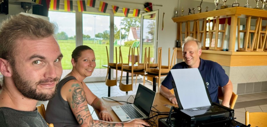 Eine Frau und ein Mann, Mitarbeiter der Gemeinde Stemwede, sitzen an einem Tisch in einem Vereinsheim und bearbeiten an einem Laptop ein Anliegen eines Bürgers, der ihnen gegenüber sitzt und für das Fot freundlich in die Kamera lacht.