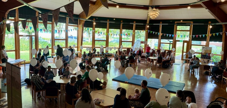 Eine Veranstaltungshalle, in der zahlreiche Erwachsene mit kleinen Kindern an Tischen sitzen. Die Halle ist mit Luftballons und Girlanden geschmückt.