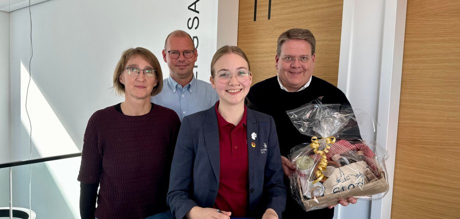 Eine junge Frau mit Brille und einem dunklen Blazer steht an einem Stehtisch und trägt sich in ein vor ihr liegendes Gästebuch ein. Hinter Ihr stehen die Eltern der jungen Frau sowie der Bürgermeister der Gemeinde Stemwede, der ein Präsent in den Händen hält.