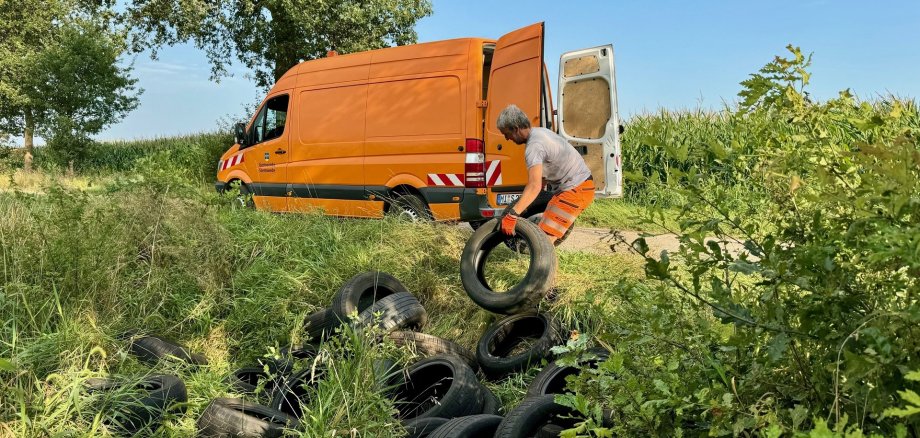 Ein Bauhofmitarbeiter der Gemeinde Stemwede ist dabei, illegal entsorgte Autoreifen aus dem Graben zu ziehen, um sie in einen orangen Bulli zu laden.