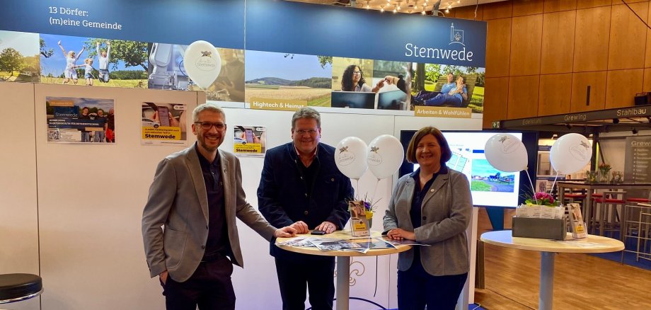 Ein Messestand, der augenscheinlich zur Gemeinde Stemwede gehört. Zwei Männer und eine Frau stehen an einem Stehtisch und lächeln in die Kamera