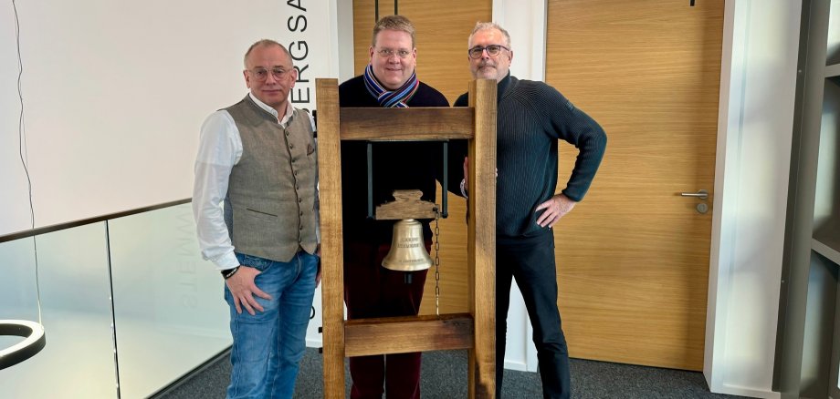 Drei Männer stehen hinter einem massiven Holzgestell, dass eine ca. 30 Zentimeter große, goldene Glocke hält. 