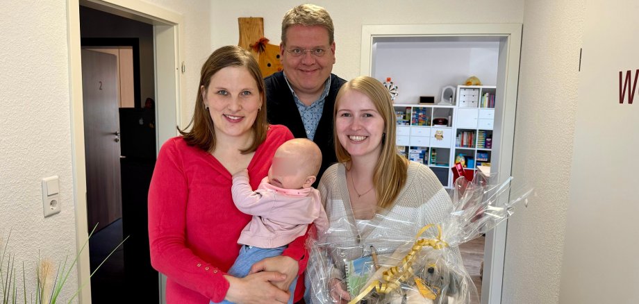 Zwei junge Frauen - die eine trägt ein Baby auf dem Arm, die andere hält ein Geschenk in den Händen - lächeln in die Kamera. Im Hintergrund steht Stemwedes Bürgermeister.