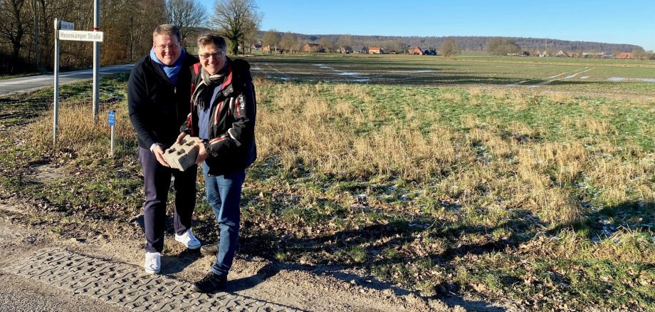 Zwei Männer stehen an einer Straße und halten exemplarisch einen Rasengitterstein, der am Wegesrand verbaut wird. 