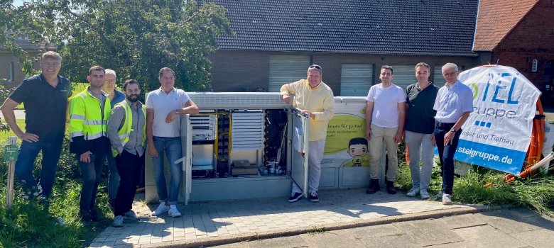 Neun Männer stehen an einem sommerlichen und sonnigen Tag vor einem Schaltkasten an einer Straße und lächeln in die Kamera. In dem Schaltkasten sind verschiedene technische Elemente zu erkennen, die für einen Glasfaseranschluss nötig sind.