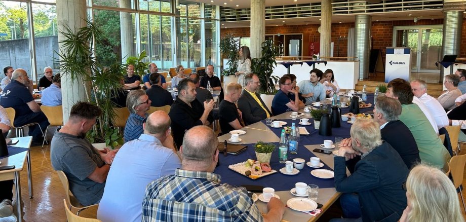 Etwa 50 Personen, die in einer Betriebskantine an verschiedenen, eingedeckten Frühstückstafeln sitzen und einer Person, die im Hintergrund steht, zuhören.