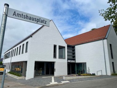 Ein Straßenschild, mit der Aufschrift "Amtshausplatz" vor einem modernen, weißen Gebäude, an dem die Aufschrift "Amtshaus" zu lesen ist.