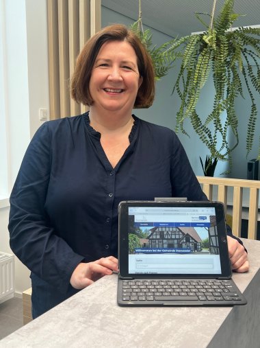 Eine Frau mit braunen Haaren und einem blauen Oberteil steht an einem Stehtisch und lächelt in die Kamera. Vor ihr steht ein geöffneter Laptop.